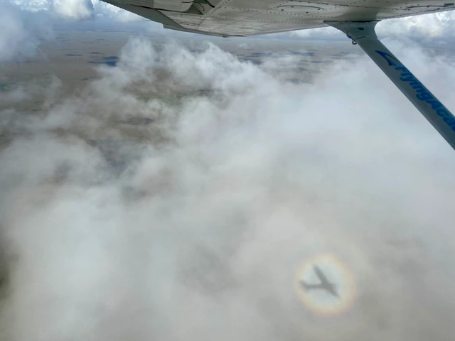 An aircraft shadow surrounded by a circular rainbow on the clouds below a wing.